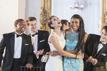 A group of five multi-ethnic teenagers and  young adults dressed in formalwear - dresses and tuxedos attending a party. The two girls are hugging each other as the men applaud.